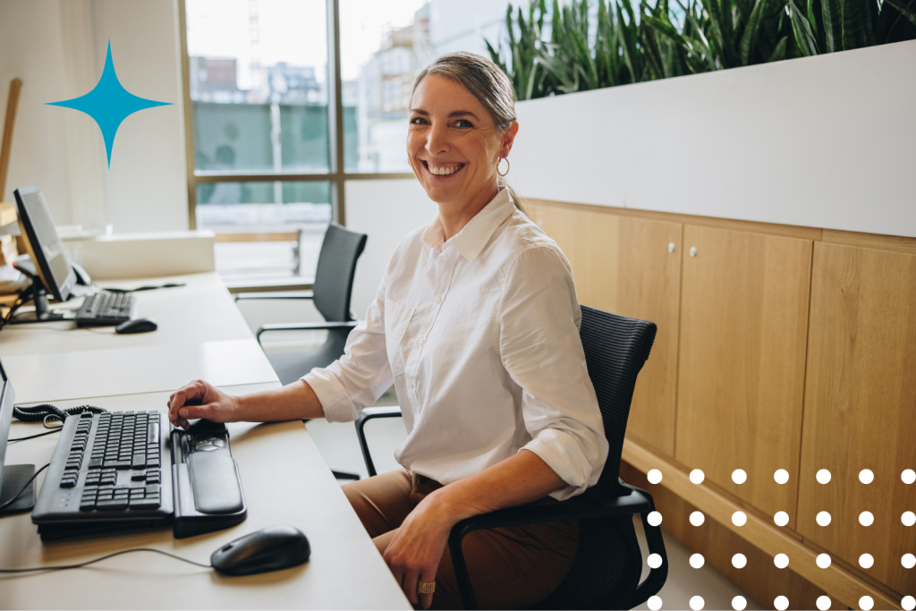Professional woman smiling at her desk in a modern office setting, manages and reports employee call-outs with Call Experts through Abridged Technology’s Attendance Bridge, creating a fully automated call flow to handle.