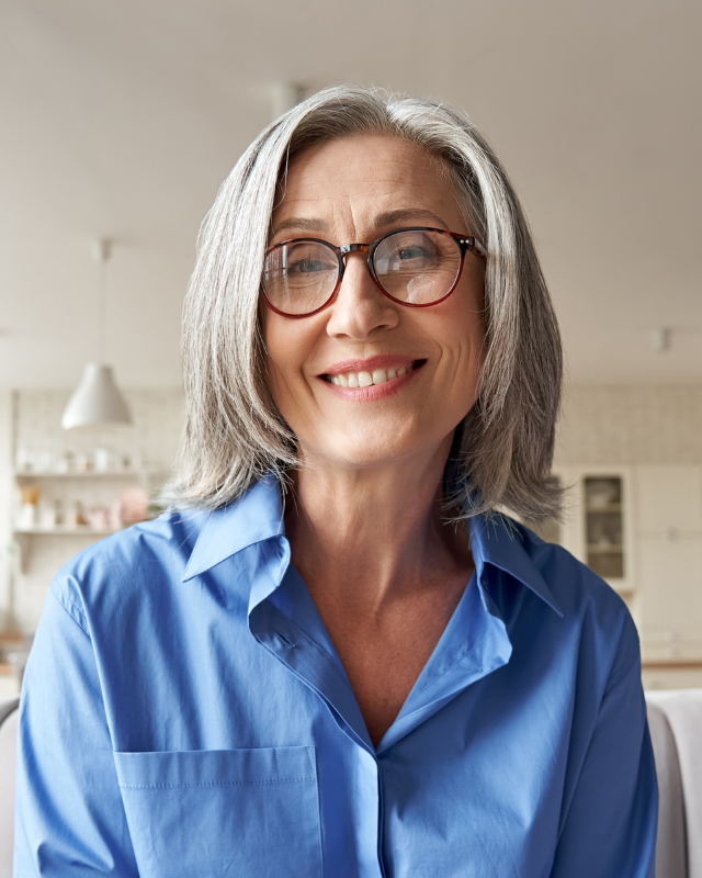 A smiling woman with gray hair and glasses wearing a blue shirt, radiating confidence and approachability in a bright, indoor setting.